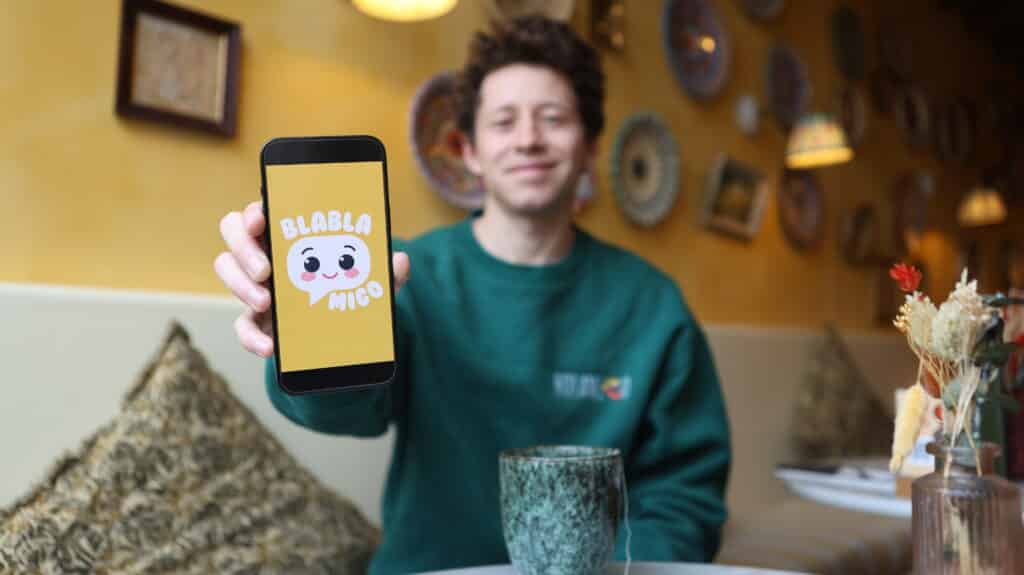 Photo de Pierre portant un sweat avec le logo de Holamigo et tenant un smartphone qui montre le logo de l'application « Blablamigo »